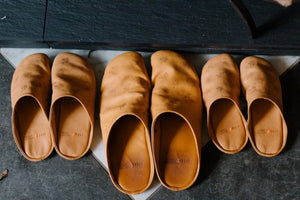 Custom-made footwear in tan leather slippers with embossed patterns arranged in three different sizes on a dark floor.