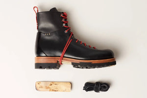 Hand sewn shoes in black leather with red laces, rugged sole, and wooden midsole, displayed with accessories