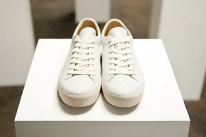 Handcrafted sneakers displayed on a white pedestal with minimalist white leather design and neat stitching.