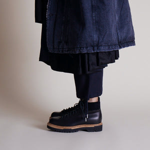 Black utility boots featuring thick tread soles and tan midsoles worn with cuffed dark pants.