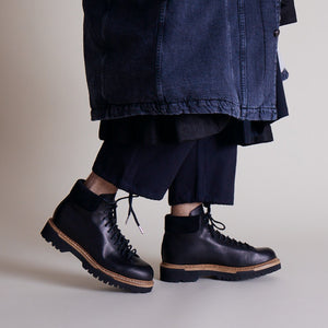 Handmade black utility boots with rugged soles and tan midsoles styled under layered dark clothing.