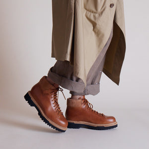 Handmade tan leather boots with black lug soles styled with rolled-up pants and a beige coat.