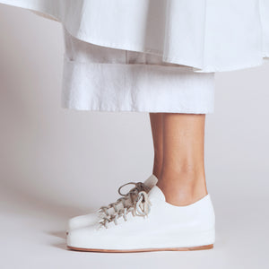 Handmade white leather sneakers worn with a mid-length white skirt and visible stitching.