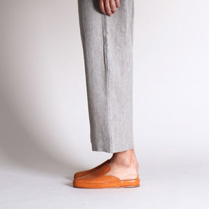 Handmade leather mules in tan worn with light gray trousers in a minimal studio setting.